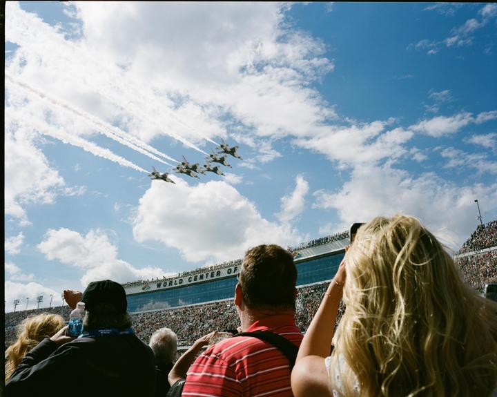 A crowd of people watches an airshow with several jets flying in formation and leaving smoke trails in the sky.