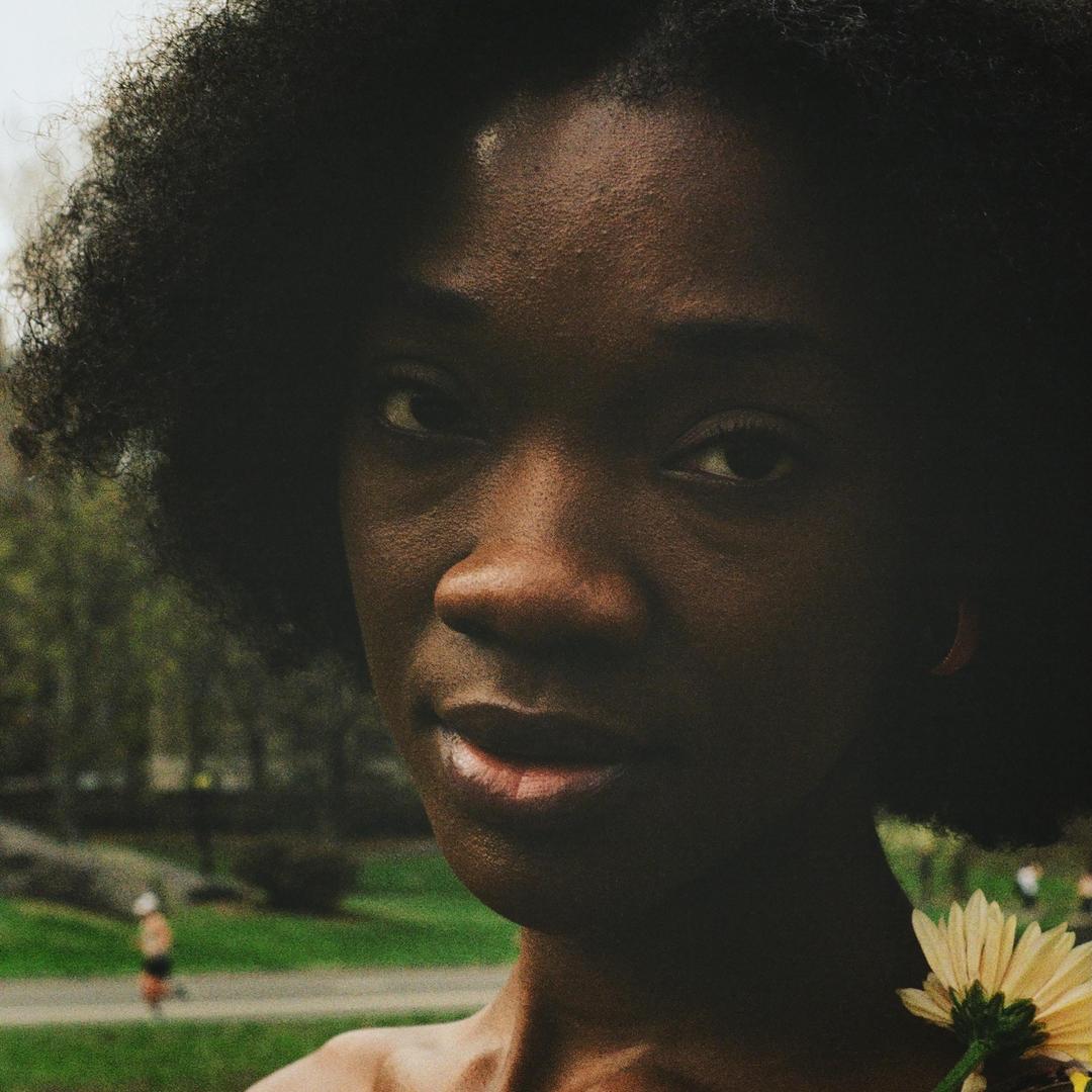 A woman with dark, curly hair looks at the viewer, holding a yellow flower on her shoulder.
