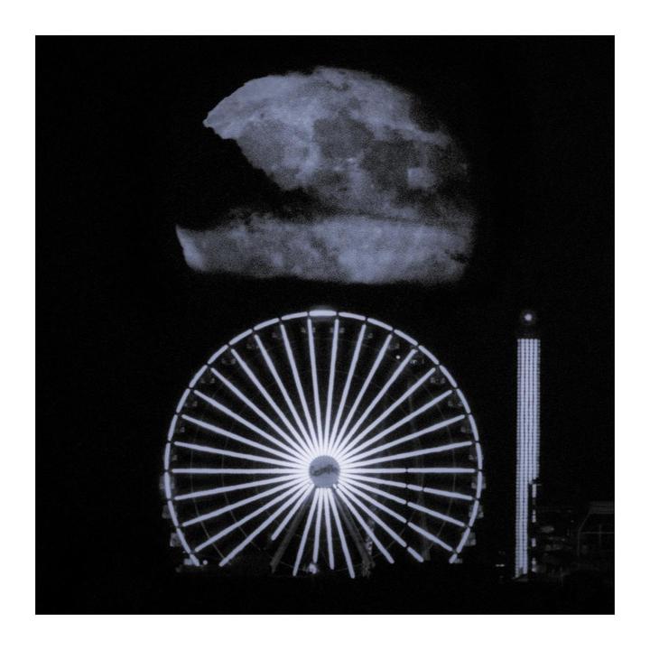 A large moon hangs above a brightly lit ferris wheel and a tall amusement park ride at night.