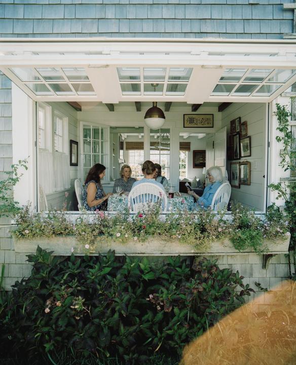 Five women are seated around a table on a porch with a window box full of flowers.