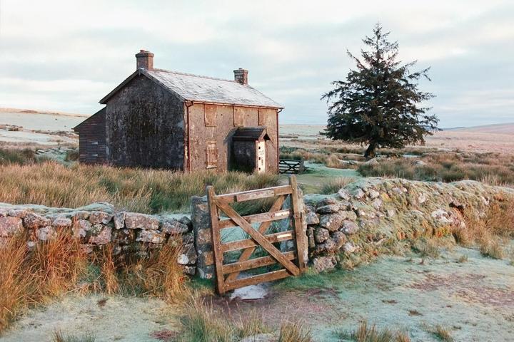 An abandoned house stands in a frosty, grassy field next to a stone wall and a wooden gate.