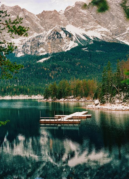 A wooden dock floats on a lake surrounded by a forest and snow-capped mountains.