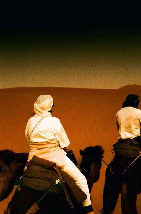 Two people ride camels across a desert landscape with a dark sky.