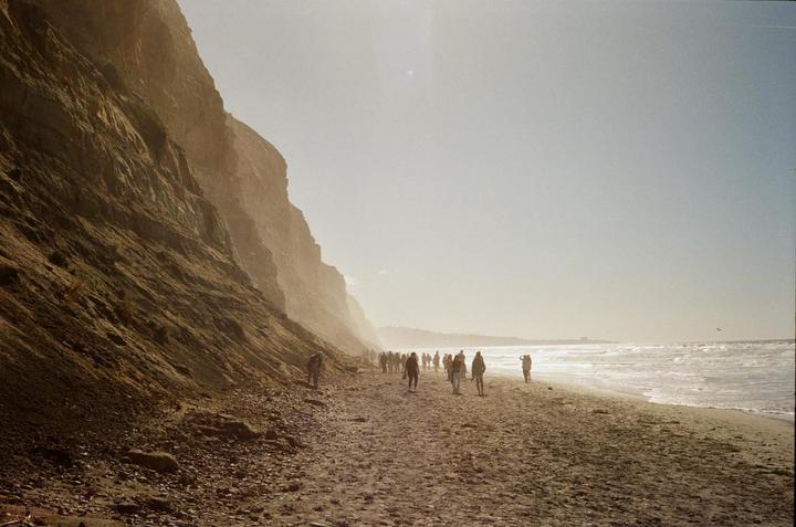 A group of people walk along a sandy beach next to towering cliffs and the ocean.