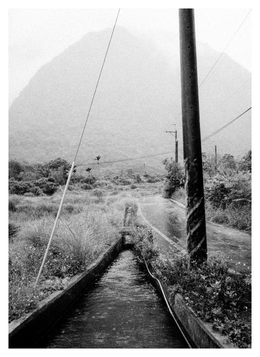 A narrow road and a water channel lead towards a hazy mountain in a rural landscape.