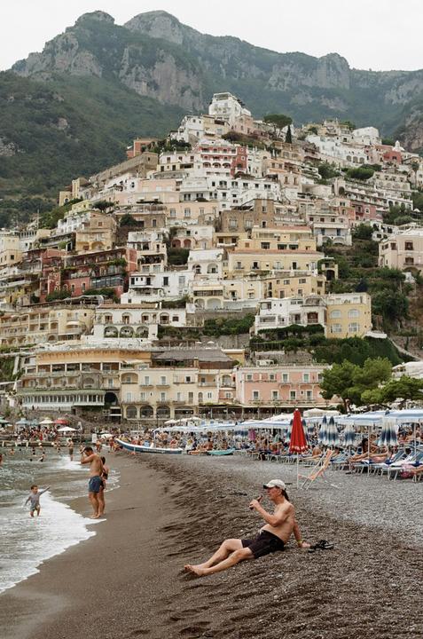 A man sits on a pebble beach with a town built into a mountain in the background.