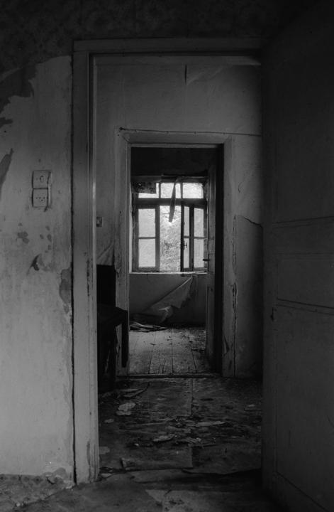 A dark, abandoned room with peeling paint on the walls and a bright window at the end of a hallway.