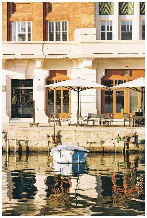 A small boat is moored in the water in front of a building with outdoor seating and umbrellas.