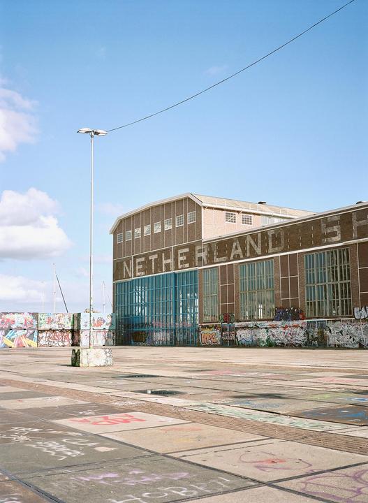 A large brick building with the word "NETHERLANDS" written on its side stands next to a concrete area covered in graffiti under a blue sky.