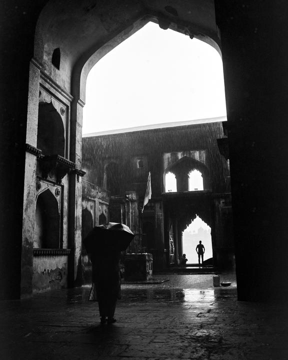 A person with an umbrella stands in a stone archway looking out at a rainy courtyard with another person silhouetted in a distant archway.