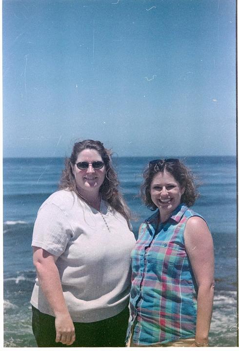 Two women stand smiling for a photo with the ocean in the background.