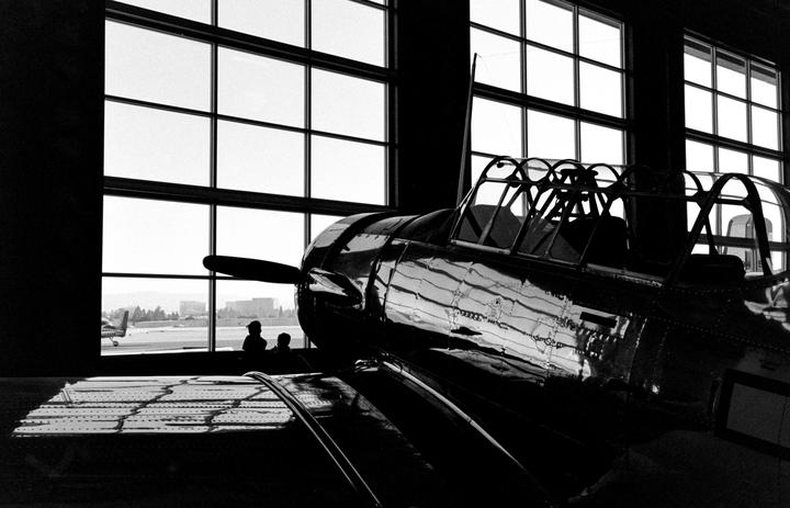 A shiny airplane is parked inside a hangar with large windows looking out onto an airfield.