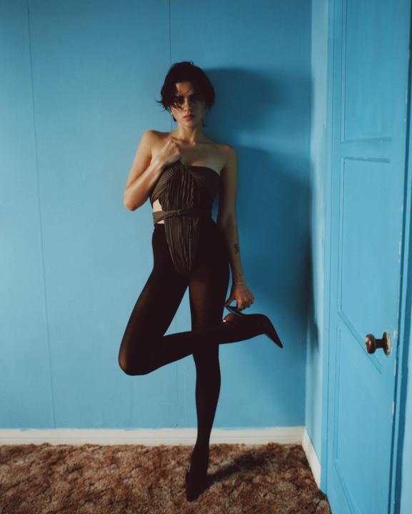 A woman in a dark outfit and tights poses against a blue wall, holding a high heel shoe.