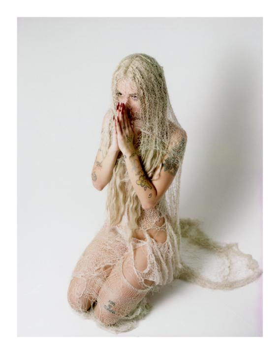 A woman with long blonde hair and tattoos kneels while wearing a tattered, net-like garment that covers her head and body.