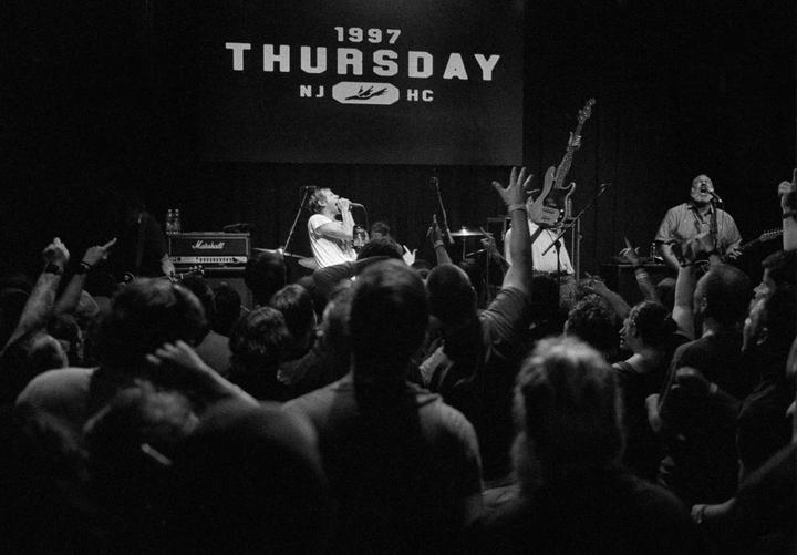 A band performs on stage in front of a crowd with a banner that reads "1997 THURSDAY NJ HC" in the background.