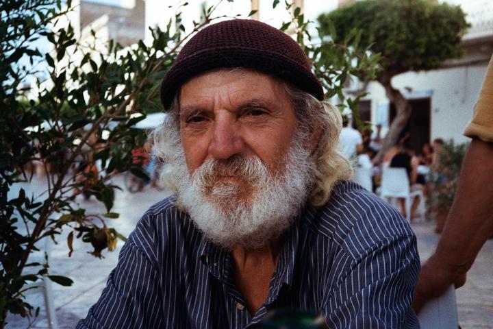 A man with a white beard and a dark knit cap looks directly at the viewer from behind some foliage.