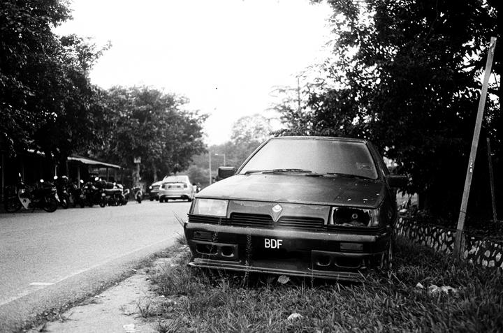 A black and white street view shows a car with a missing headlight parked on a grassy verge next to a road with motorcycles and another car in the background.