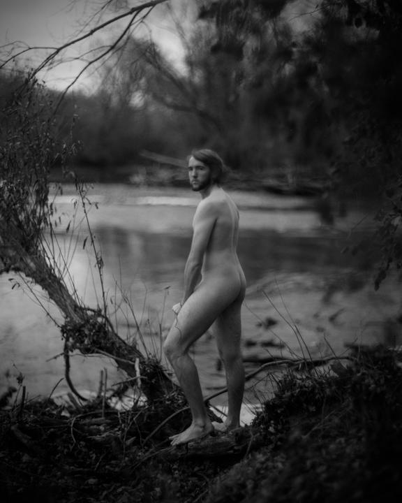 A man stands nude on a riverbank, looking over his shoulder.