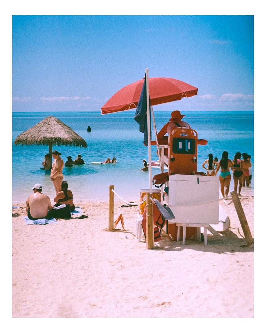 A lifeguard sits under a red umbrella on a beach with people swimming in the ocean.