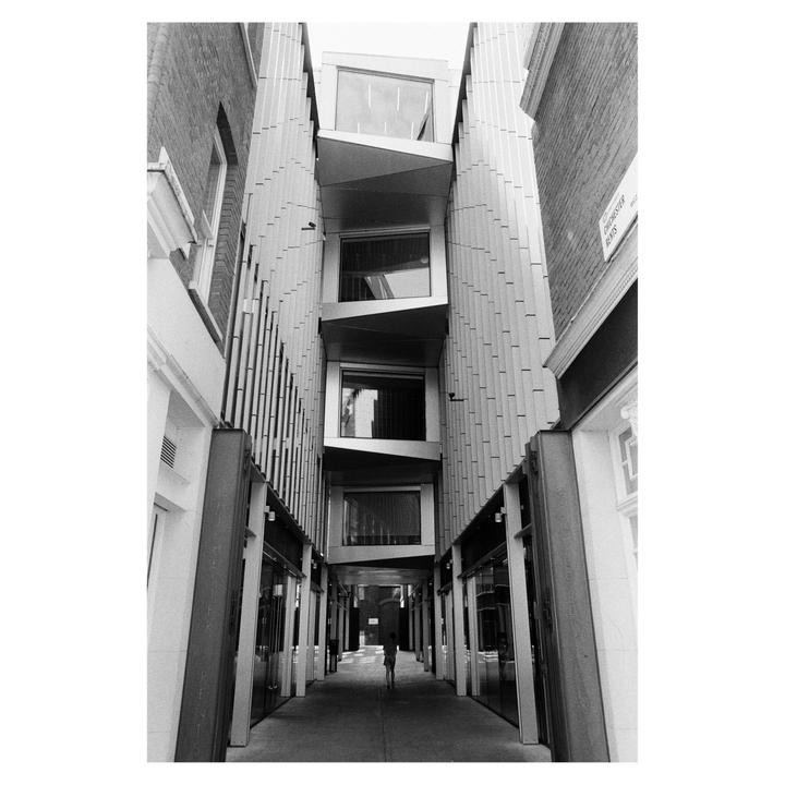 A person walks through a narrow alleyway between two tall buildings, one with a brick facade and the other with a modern, angular design.