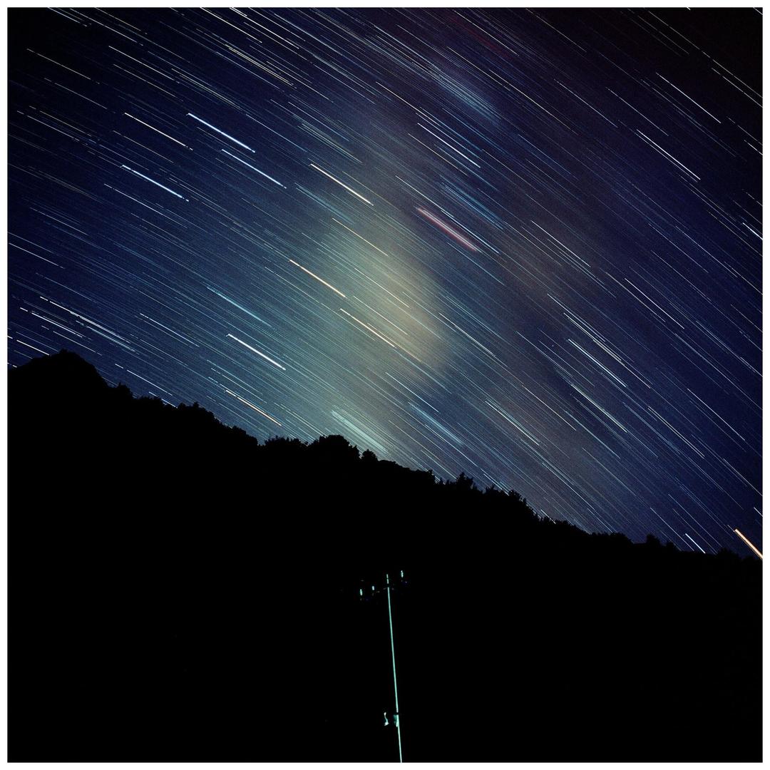 A long exposure photograph captures star trails across a night sky above a dark mountain silhouette and a utility pole.