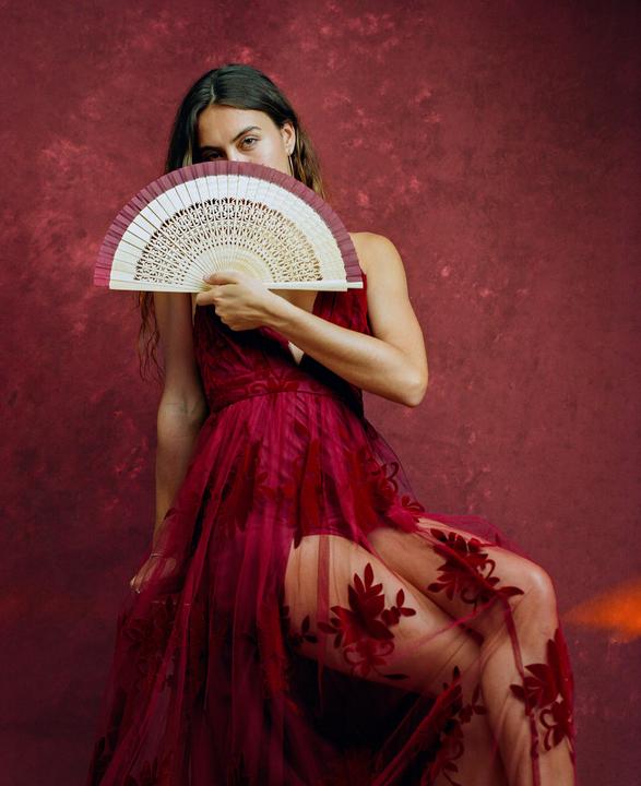 A woman in a red dress holds a fan in front of her face against a red background.