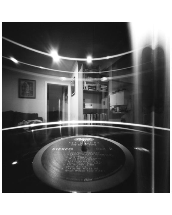 A record player with a vinyl record is in the foreground, with a room and bookshelves visible in the background.