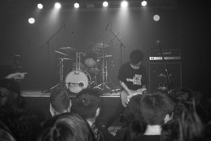 A musician plays guitar on a stage with a drum kit and amplifiers in the background, viewed from the audience.