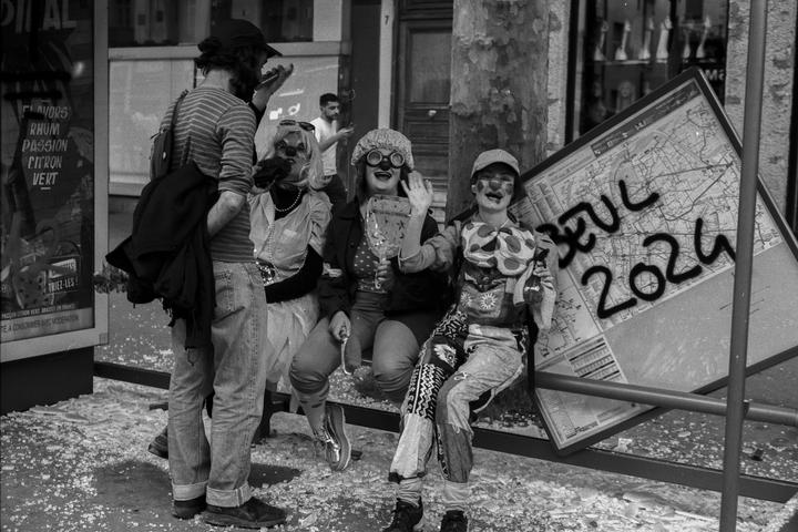 Three women in clown makeup and costumes sit on a bench next to a man on a street covered in confetti.