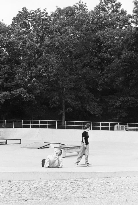 A woman on a skateboard and another woman sitting on the ground are in a skatepark surrounded by trees.