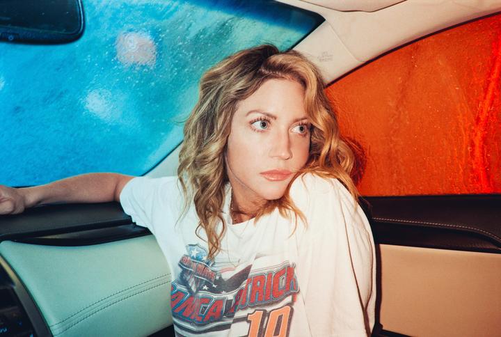 A woman with blonde hair sits in a car with blue and orange light reflecting on the windows.