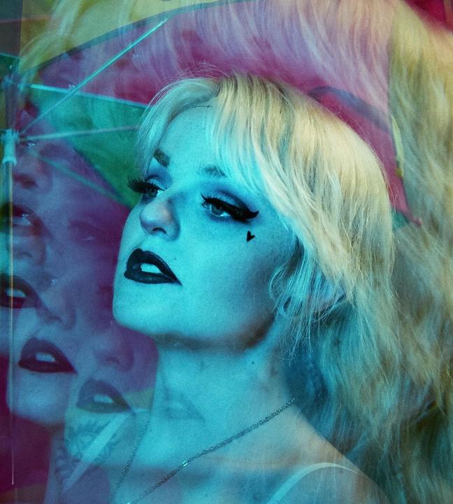 A woman with blonde hair and dark lipstick looks up and to the right, with multiple exposures of her face and an umbrella visible in the background.