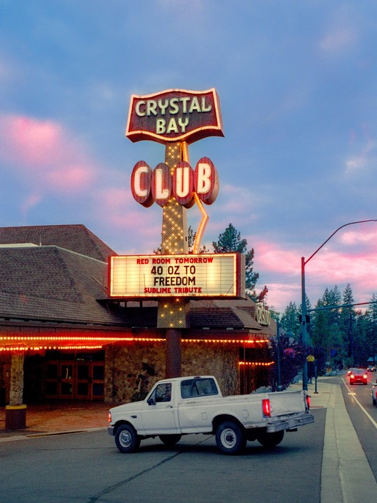 A white pickup truck is parked in front of the Crystal Bay Club with its neon sign illuminated against a dusk sky.