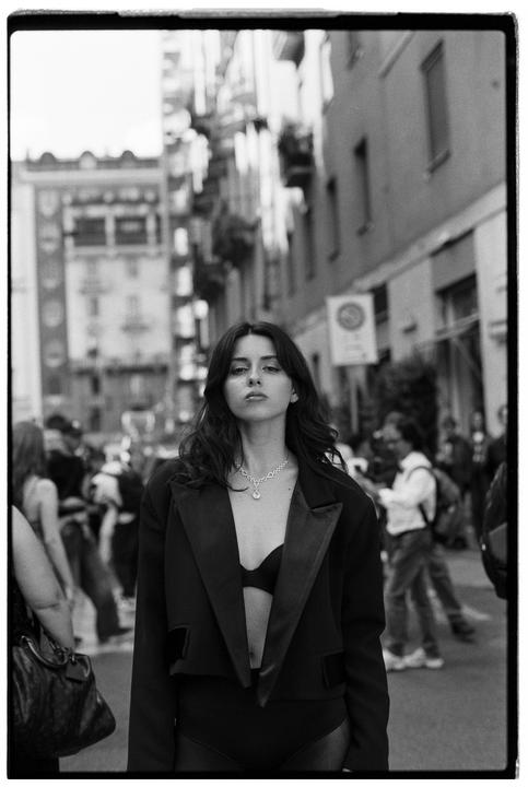 A woman wearing a blazer and bra stands on a city street.