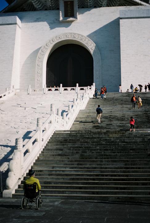 A person in a wheelchair looks up at a grand white building with a large staircase and many people.