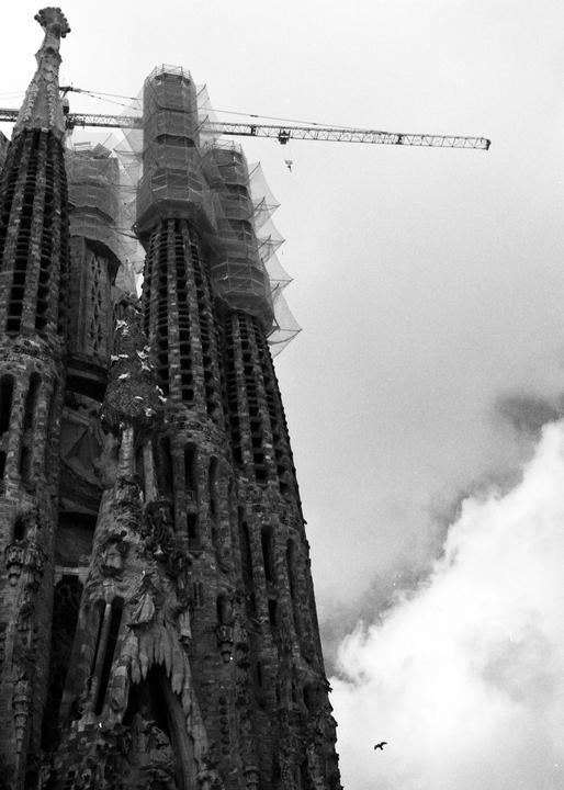 The Sagrada Familia cathedral is under construction with a crane and scaffolding visible against a cloudy sky.