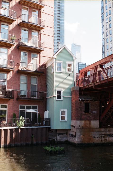 A small green house stands next to a brick building and a red bridge over a river in a city.