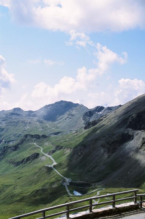 A winding road and a small lake are visible in a mountainous landscape under a cloudy sky.