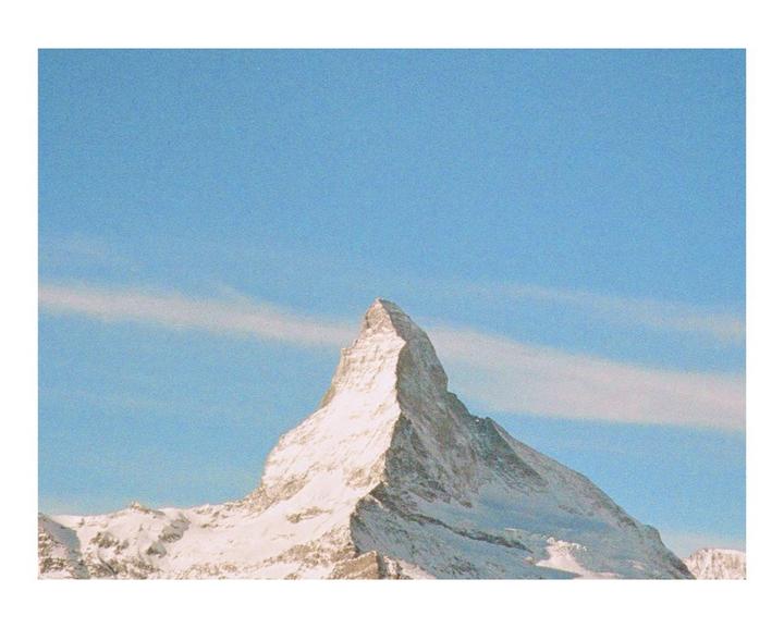 A snow-covered mountain peak rises sharply against a clear blue sky with a faint cloud.