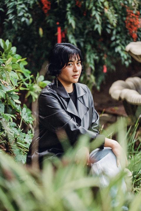 A woman wearing a black jacket and jeans is sitting among green foliage with red berries in the background.