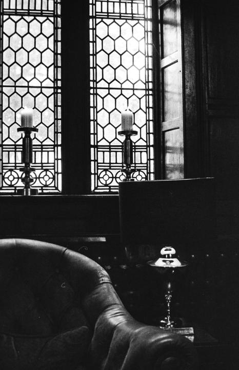 Two lit candles in ornate holders sit on a windowsill in front of a leaded glass window, with a leather armchair and a small table with a goblet in the foreground.