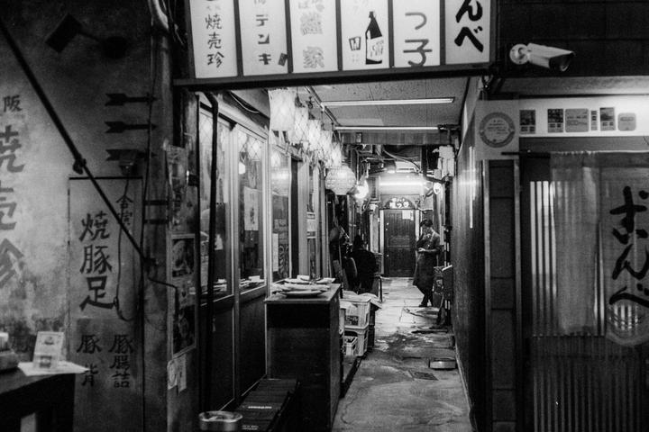 A man stands in a narrow, dimly lit alleyway lined with shops and signs.