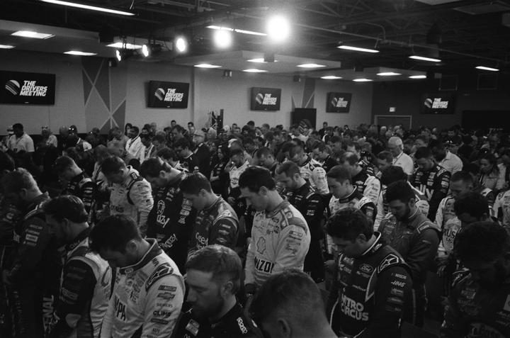 A large group of men in racing suits are gathered in a room with their heads bowed.