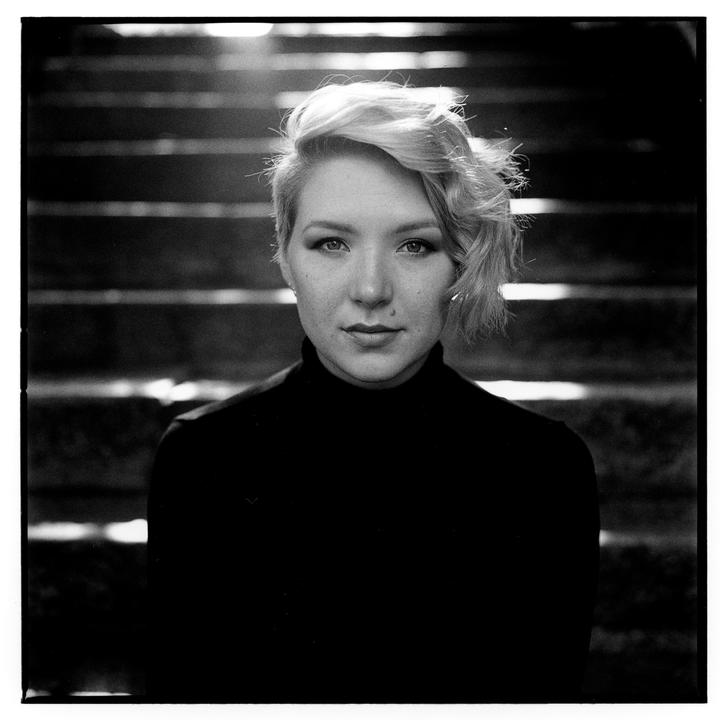 A woman with short, light-colored hair and a dark turtleneck looks directly at the viewer.