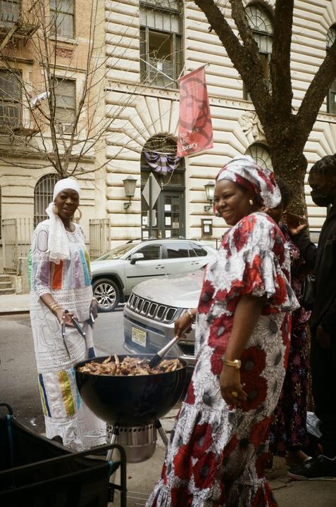 Two women in traditional clothing cook meat on a grill on a street in Harlem.