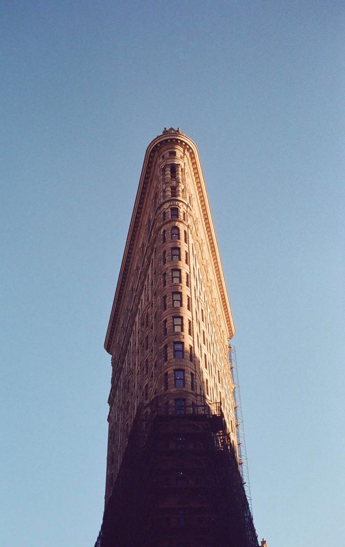 The Flatiron Building stands tall against a clear blue sky, with one side illuminated by sunlight and the other in shadow.