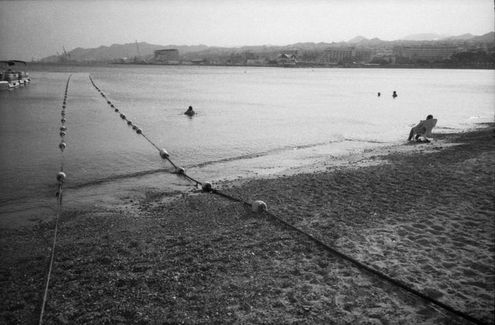 A person sits on a chair at the edge of a beach with two people swimming in the water and a city in the distance.