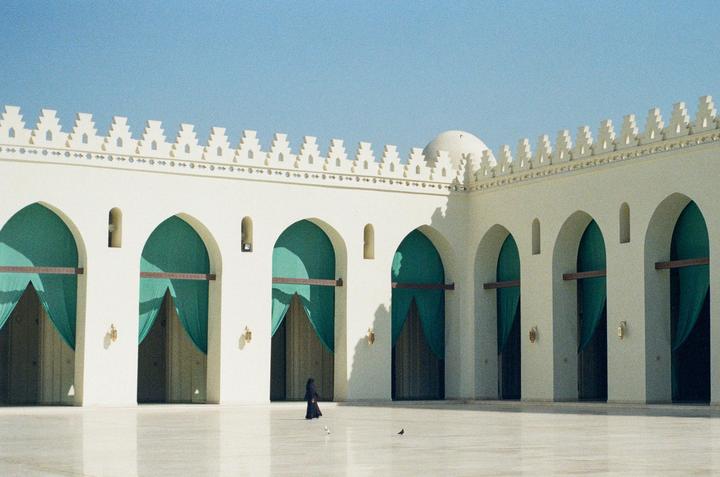A woman in a black dress walks across a large, light-colored courtyard with many arches and green curtains.