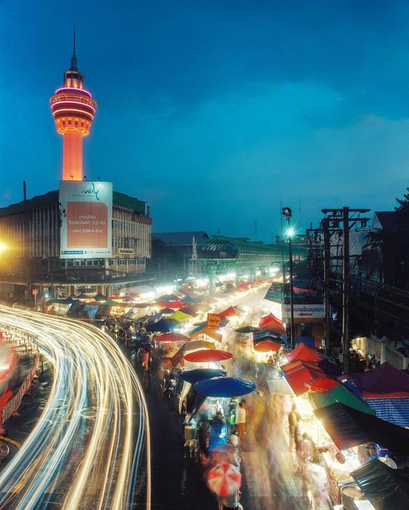 A tall, illuminated tower overlooks a busy night market with light trails from passing vehicles.