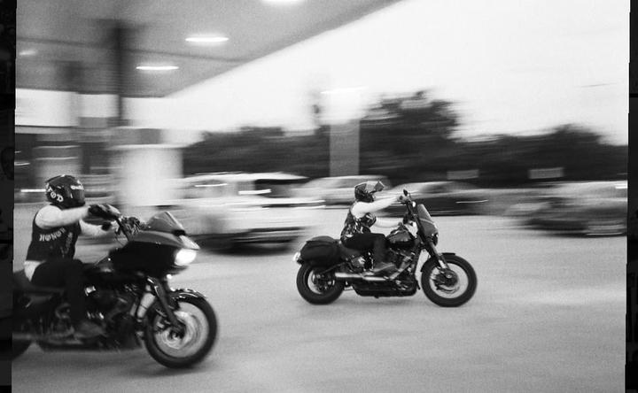 Two motorcyclists ride on a street at night, with blurred cars in the background.